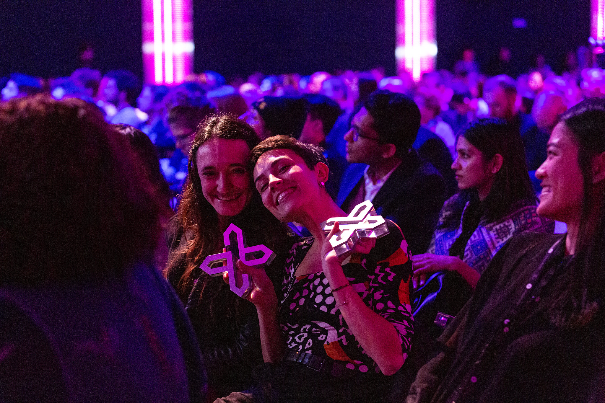 Two smiling women seated in the Interaction Awards audience hold their Awards trophies up to the camera.