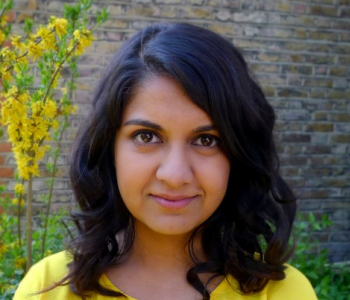 Head shot of Anab Jain looking straight at the camera. She's wearing a yellow top that matches the yellow plant behind her.